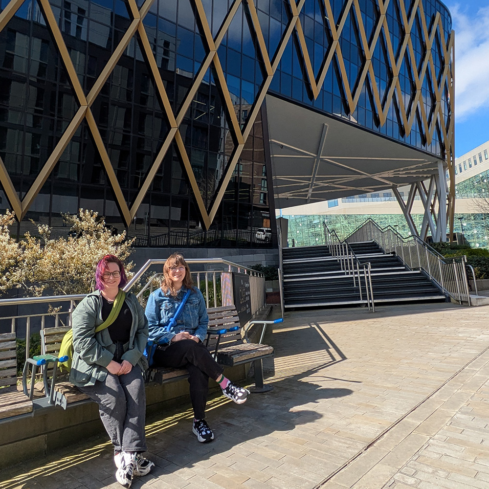 Ellen and Michelle sitting on a bench outside the Catalyst Building, which is a glass fronted building with golden criss-cross. Ellen is wearing denim and has brown hair and glasses with a blue bag, while Michelle has purple hair and is wearing black with a green bag. Both are smiling and it's a lovely sunny day.