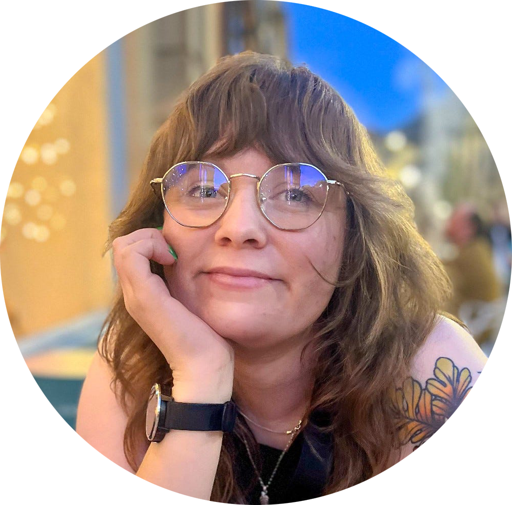 Ellen has light brown hair with layers and a fringe. She wears round, silver-rimmed glasses. She is smiling and resting her head on her hand.