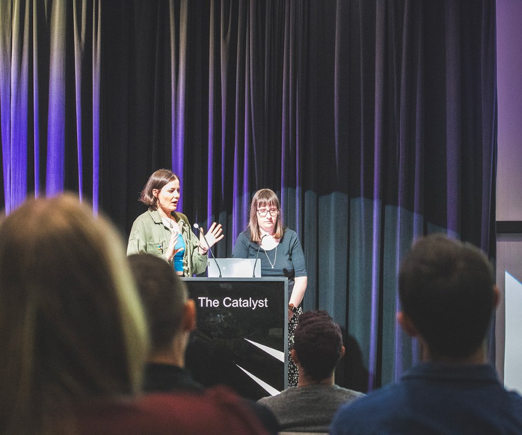 Two women, Helen and Holly, who both have brown hair. Holly has glasses. Helen, on the left, is speaking at a podium labeled “The Catalyst” in front of an audience, with stage lighting and curtains behind them.
