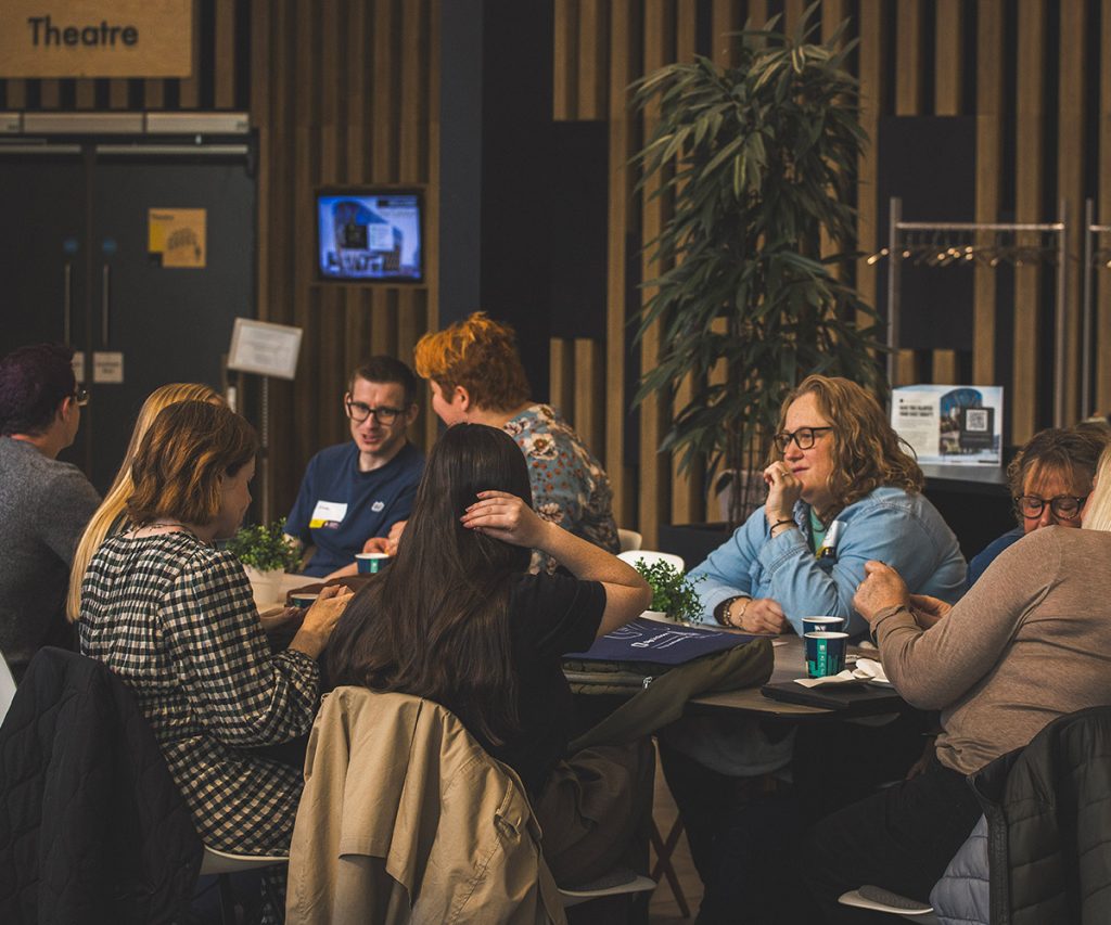 People sitting around tables in a casual discussion setting inside a modern venue, with “Theatre” signage visible in the background.