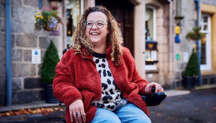 Carrie-Ann has curly hair and glasses smiling while seated in a powered wheelchair on a cobbled street, wearing a red jacket and patterned jumper, with stone buildings and shopfronts in the background.