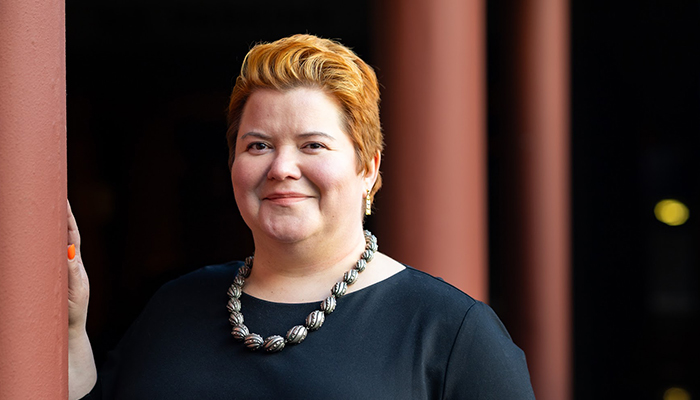 Lisa has short auburn hair and is wearing a black top and chunky silver necklace, smiling gently while standing beside a pillar in an outdoor setting.
