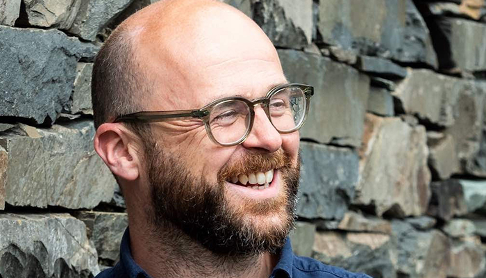 Martin is a friendly-looking white man with a bald head, short brown beard, and glasses. He’s wearing a casual dark blue shirt and is leaning against a grey stone wall.