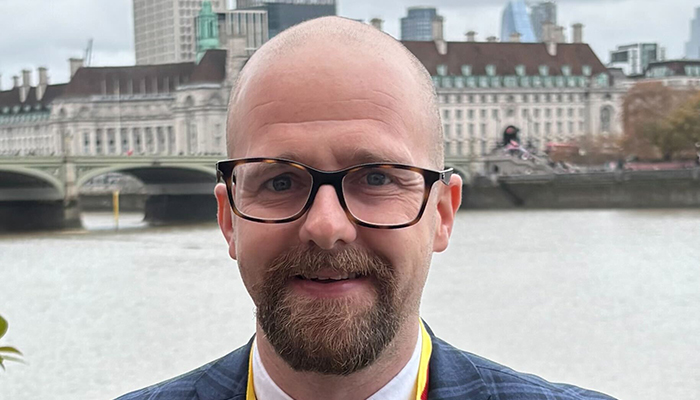Simon is a white man with glasses, and a short beard smiling at the camera, wearing a jacket and lanyard, with a river, bridge, and city buildings in the background.