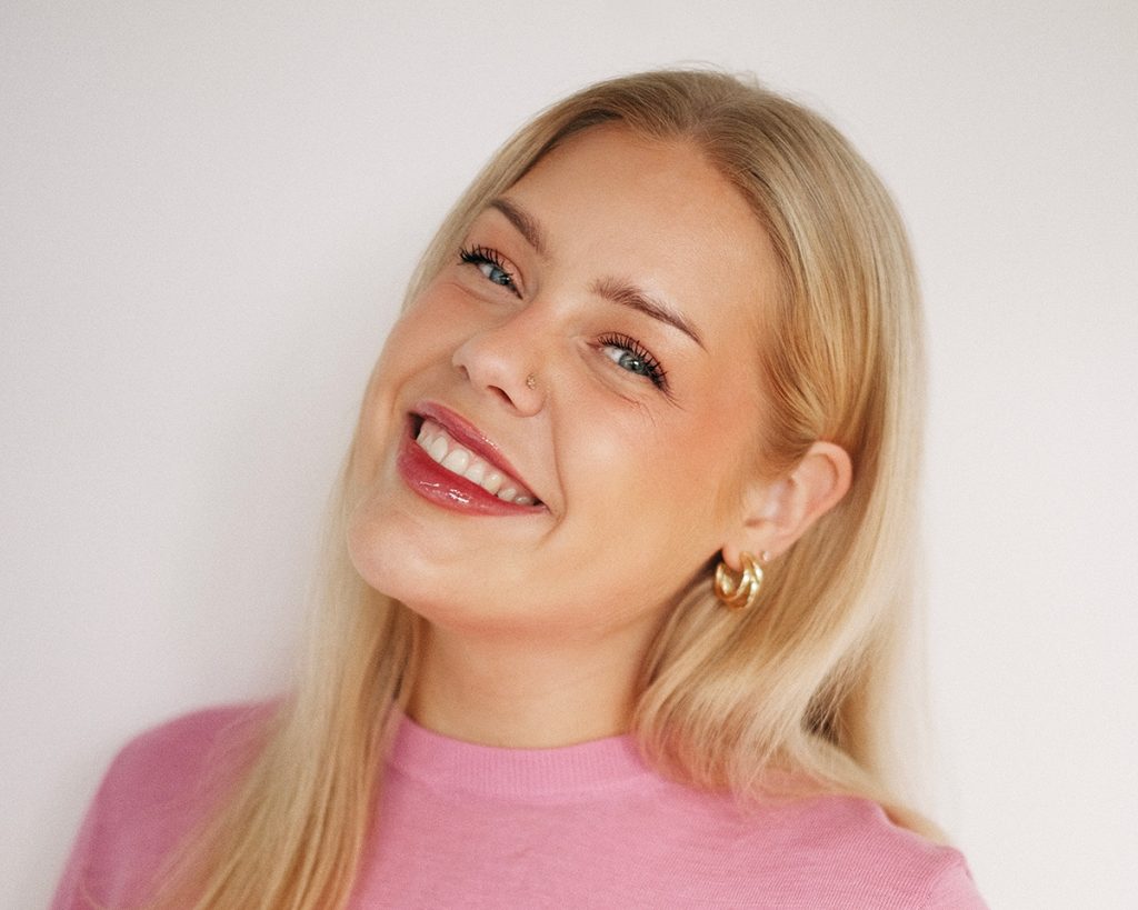 Portrait of Elza Parisa smiling with her head slightly tilted, wearing a pink top, gold hoop earrings, and a small nose stud.