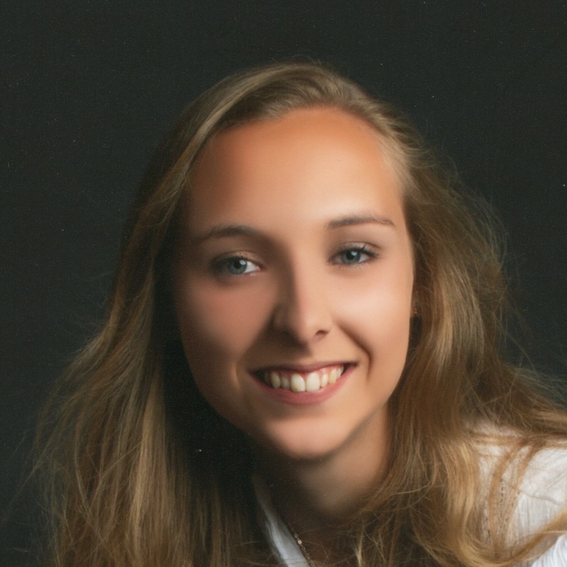 Portrait of Samantha Merrett smiling at the camera, with long light brown hair worn down and a dark top, set against a dark background.
