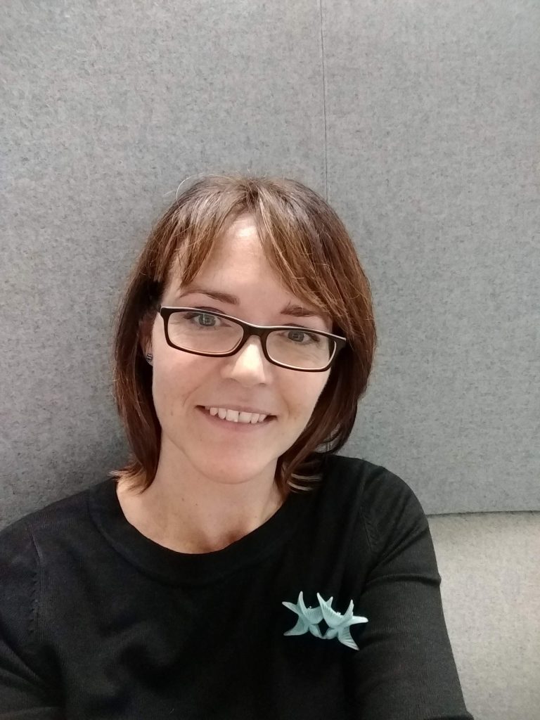 Portrait of Jane McFadyen smiling at the camera, with short brown hair and glasses, wearing a black top against a grey background