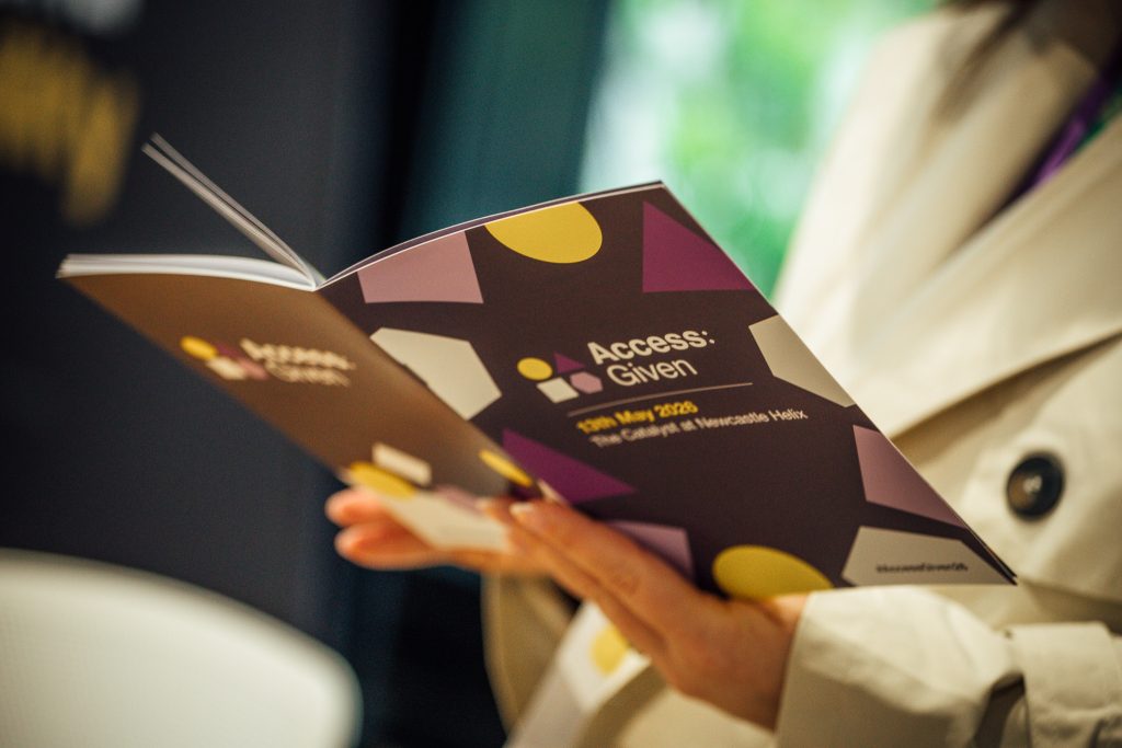 A person's hands holding a booklet for Access:Given 2026, the outer cover is purple with yellow and white shapes.